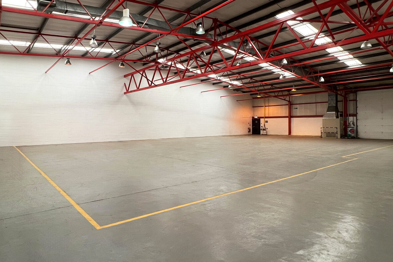 Large, empty industrial warehouse with grey concrete floors, high ceilings, red steel beams, and overhead lighting; yellow lines mark a section on the floor.
