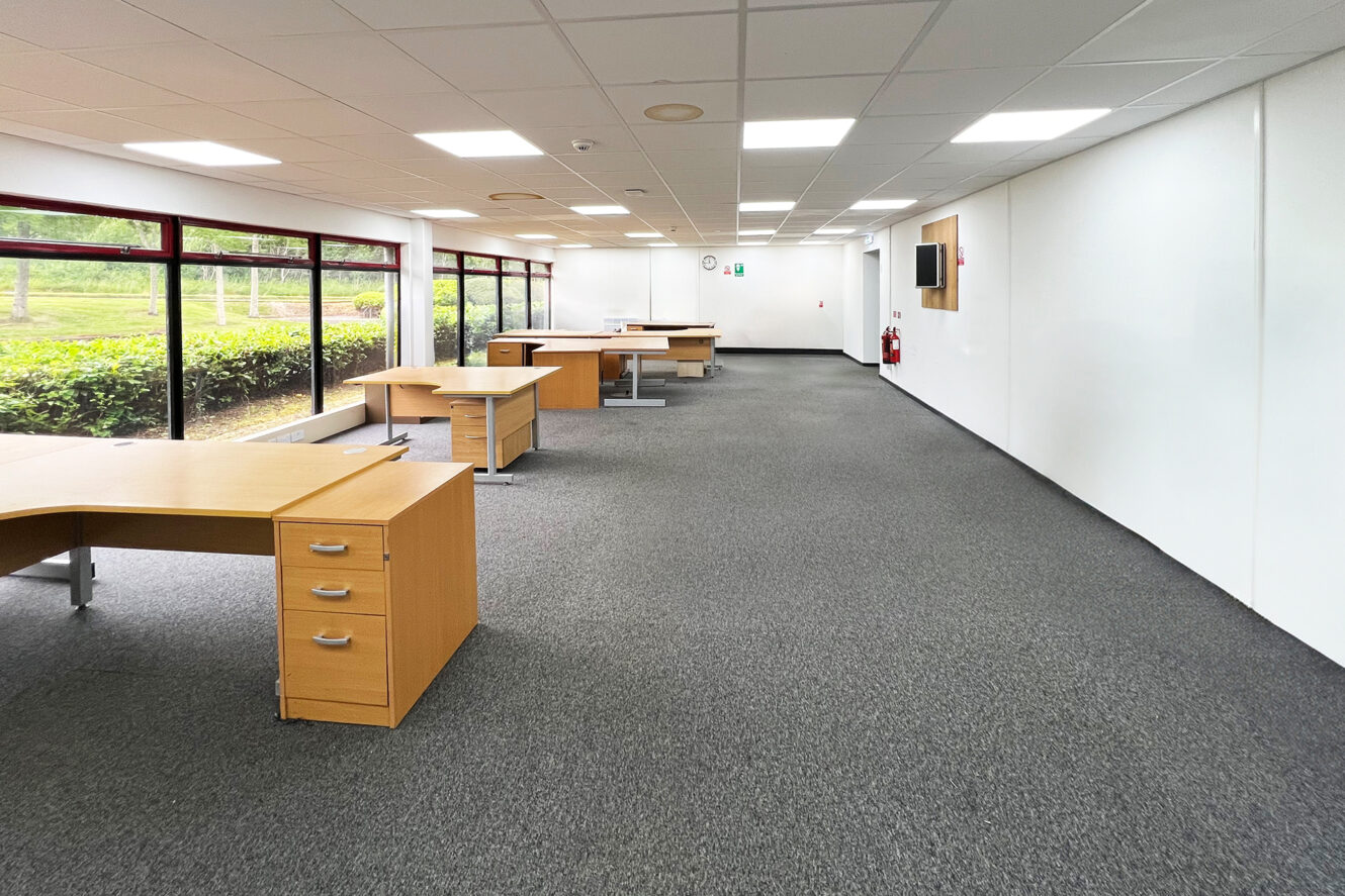 Modern office space with several empty wooden desks, large windows on one side, white walls, carpeted floor, and emergency equipment visible on the wall.