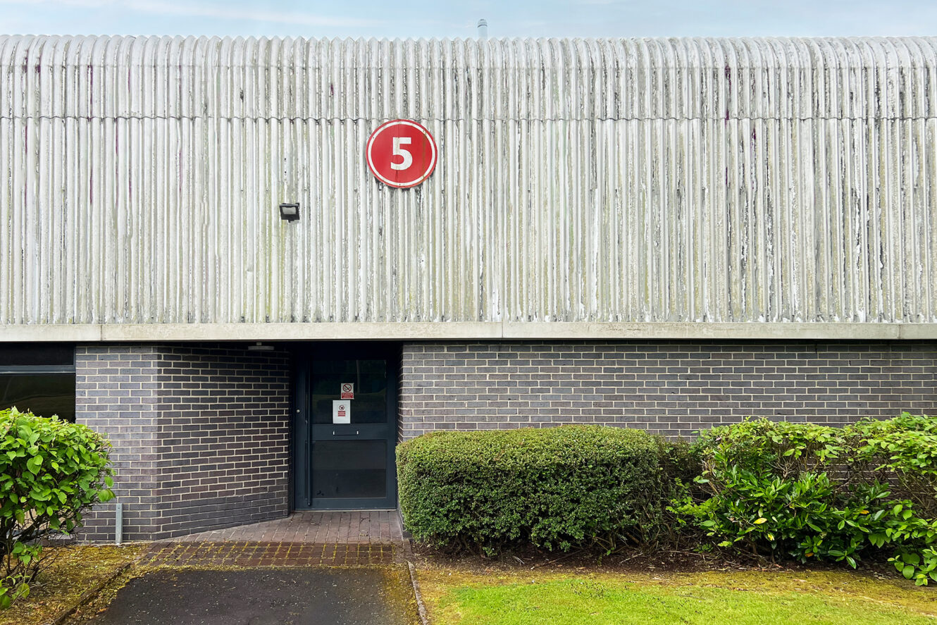 A concrete and brick building with a door and a large red number 5 sign above it, surrounded by green bushes and grass.