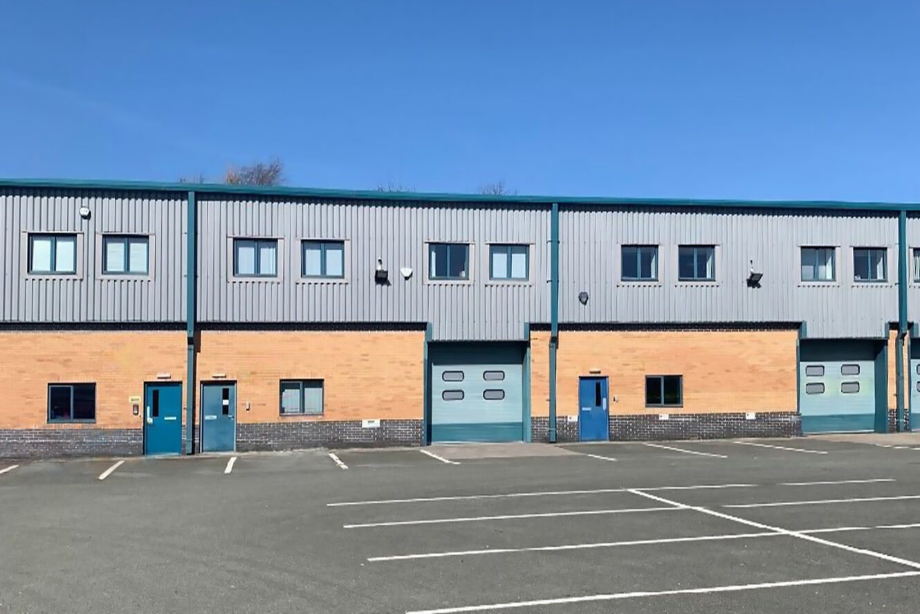 A modern industrial building with brick and metal exterior, multiple blue doors, large garage doors, and an empty parking lot in front under a clear blue sky.