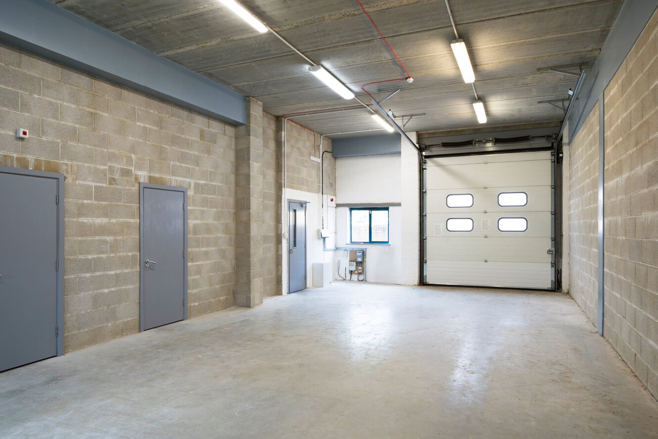 Empty industrial garage with concrete floor, cinder block walls, overhead fluorescent lights, a metal door, and a large roll-up garage door letting in natural light.
