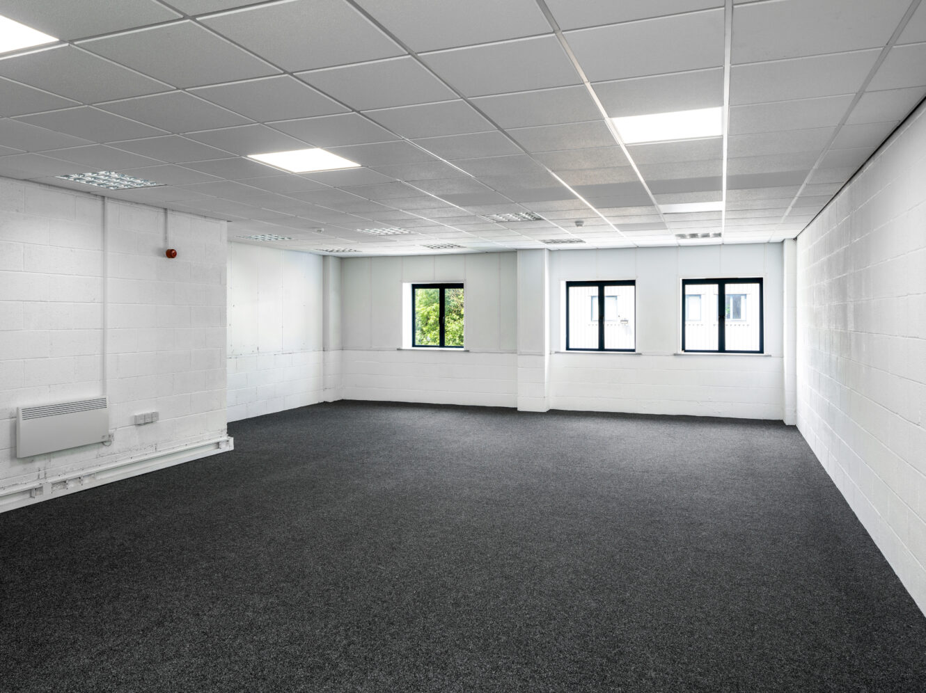 Empty office room with white walls, carpeted floor, fluorescent ceiling lights, three windows, and a wall-mounted heater.