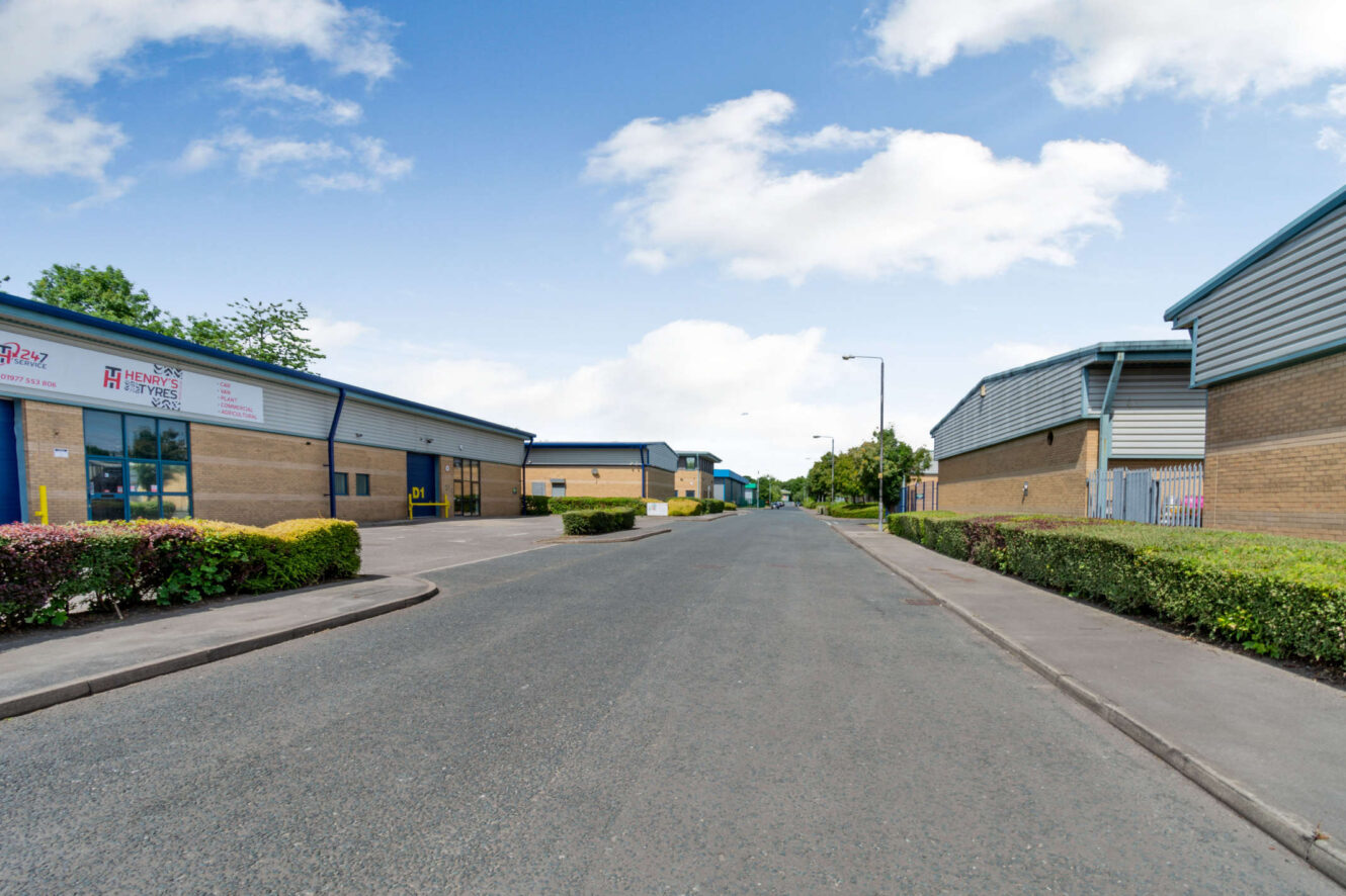Empty road flanked by modern industrial buildings with yellow-brick walls and metal roofs under a partly cloudy sky.