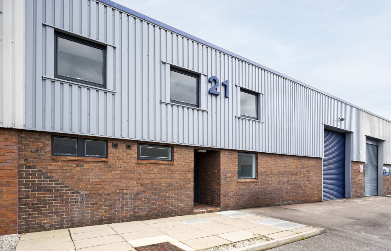 A modern industrial warehouse with brick and metal siding, three windows, a large blue roller shutter door, and the number 21 displayed above the entrance.