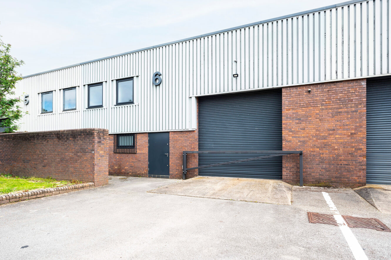 A modern industrial warehouse with a brick lower façade, gray upper panels, two garage doors, a small entrance door, and a paved parking area in front.