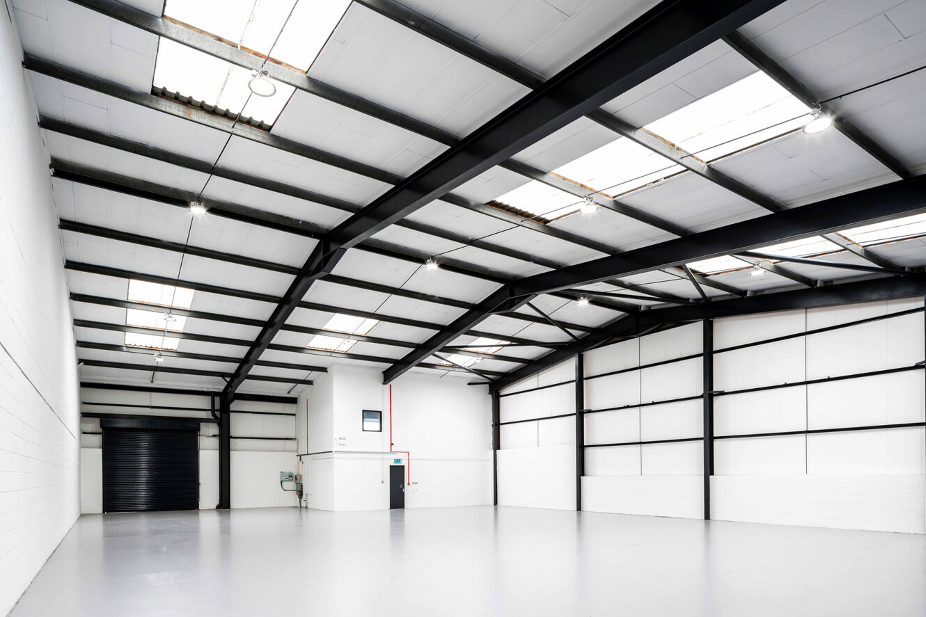 Spacious empty warehouse with white walls, high ceiling, skylights, black steel beams, and a large black roller door.