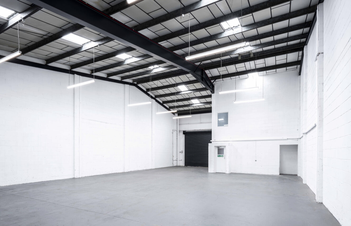 Spacious empty warehouse with white walls, high ceiling, exposed beams, fluorescent lights, and a large black roller door.