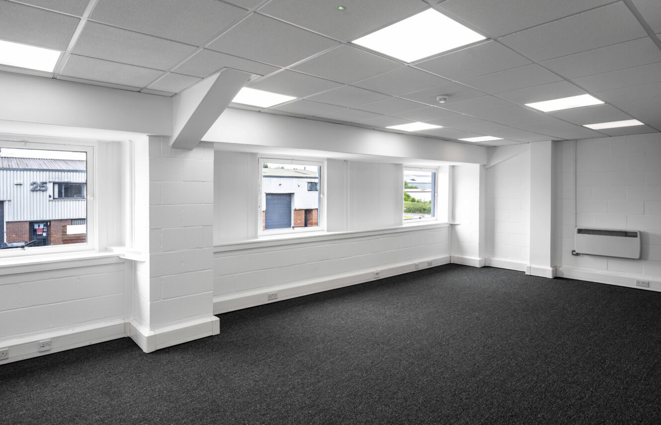 An empty, modern office space with white walls, large windows, and dark carpeted flooring. Fluorescent ceiling lights are turned on.