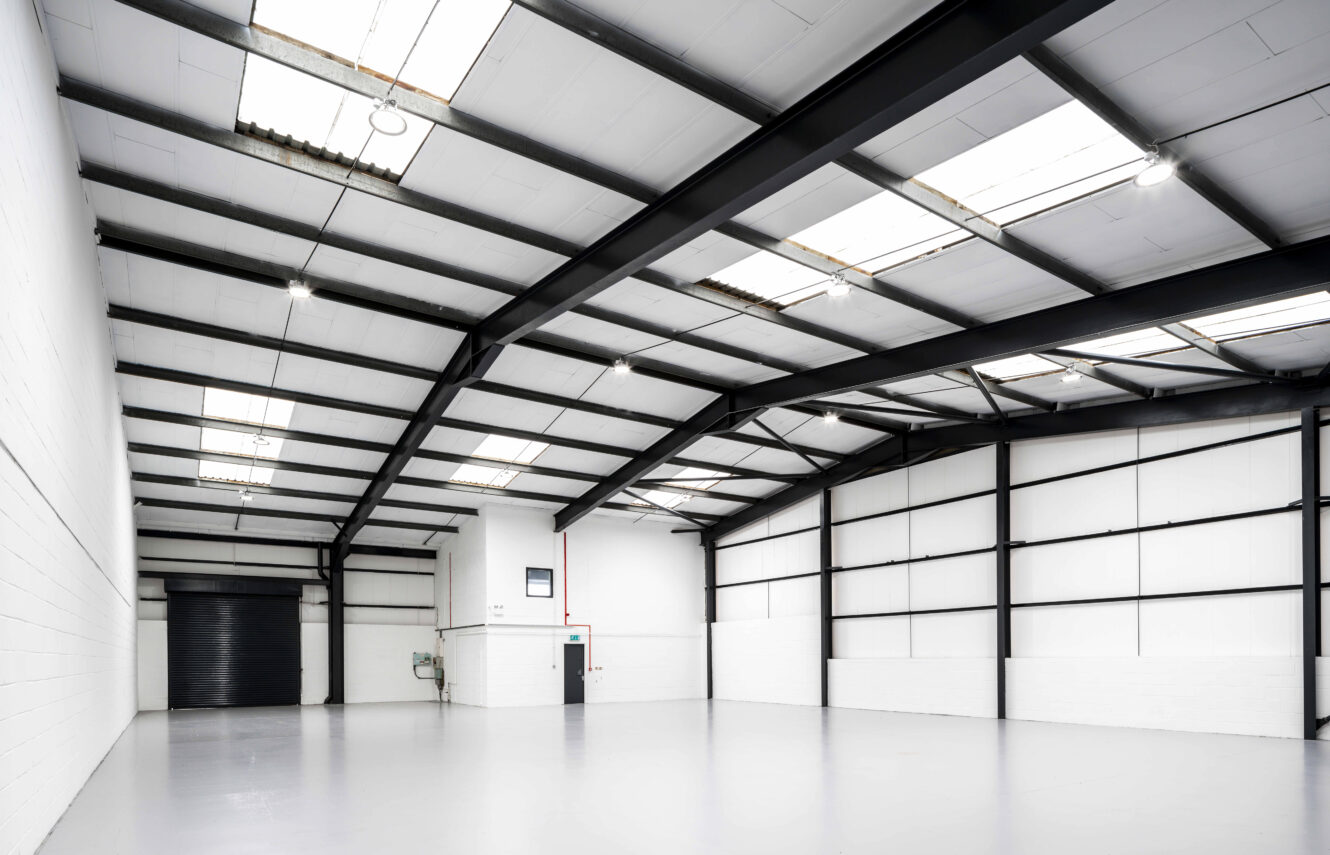Spacious, empty warehouse interior with white walls, high ceiling, exposed black beams, skylights, and a closed black roller door.
