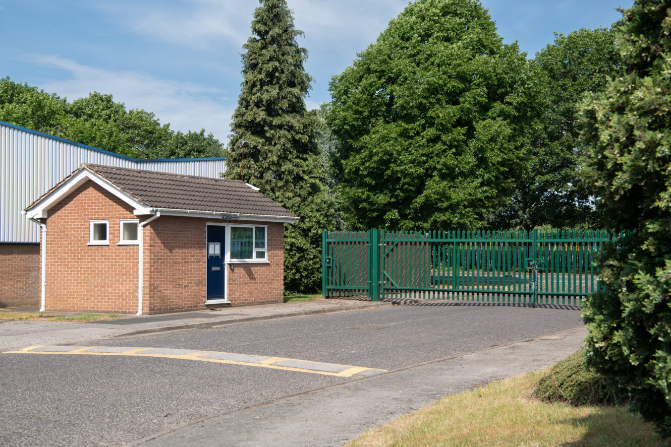 A small brick security building with a blue door stands next to a green metal gate at the entrance of a fenced property, surrounded by trees and greenery.