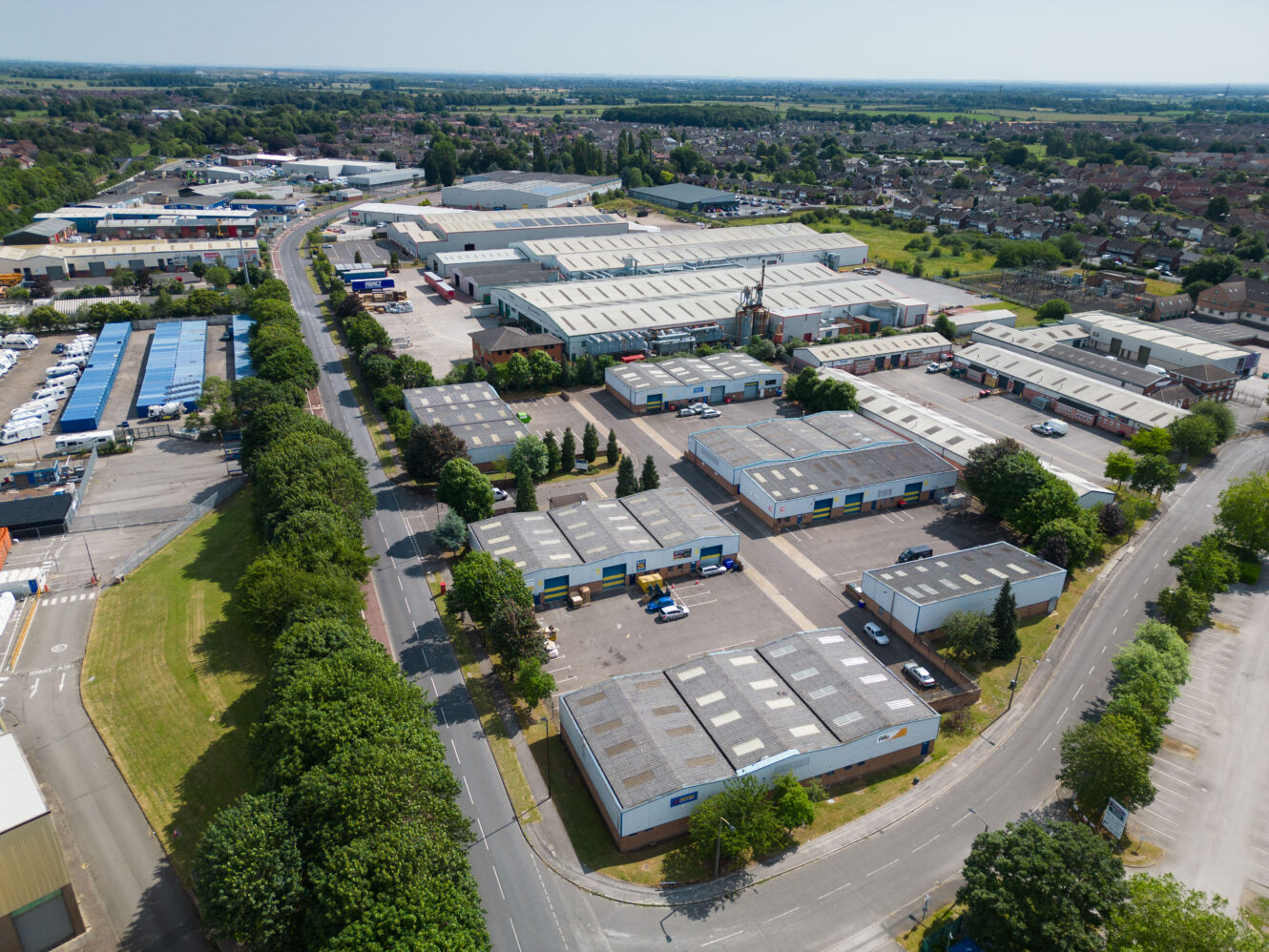 Aerial view of an industrial estate with multiple warehouses, office buildings, parked vehicles, surrounding roads, and greenery, situated on the edge of a residential area.