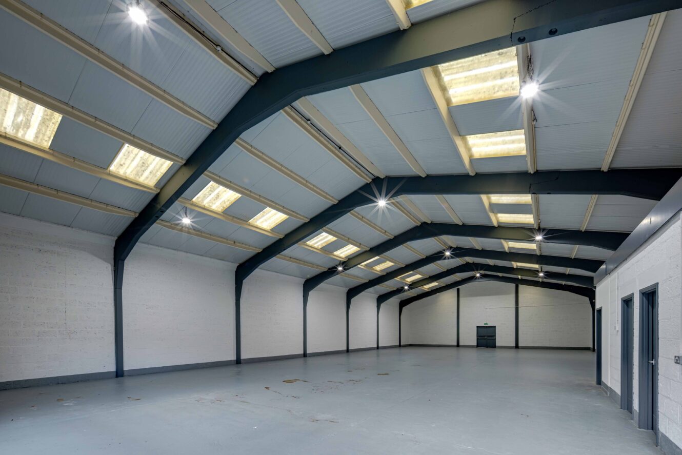 Wide-angle view of an empty industrial warehouse with gray floors, white walls, and a high ceiling with exposed beams and skylights.