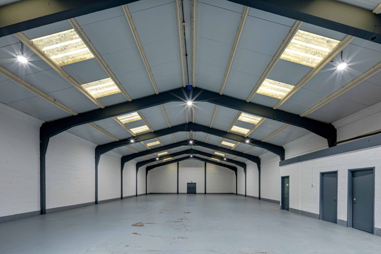 Interior of an empty industrial warehouse with high ceilings, exposed beams, multiple skylights, white walls, gray doors, and a polished concrete floor.