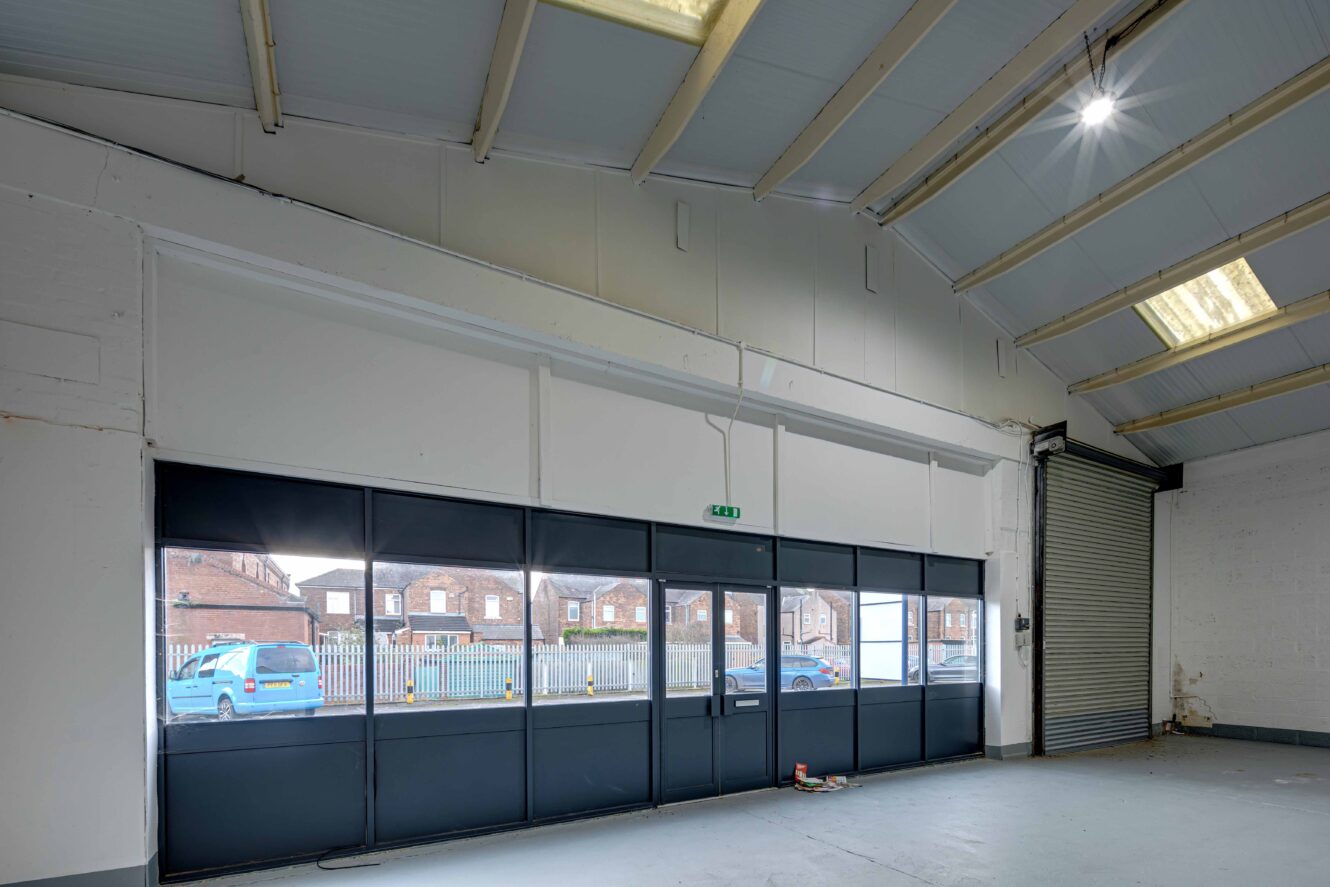 Interior of an empty warehouse with large windows, a roller shutter door, high ceiling, and visible fluorescent lighting panels. Blue car and brick buildings are visible outside.