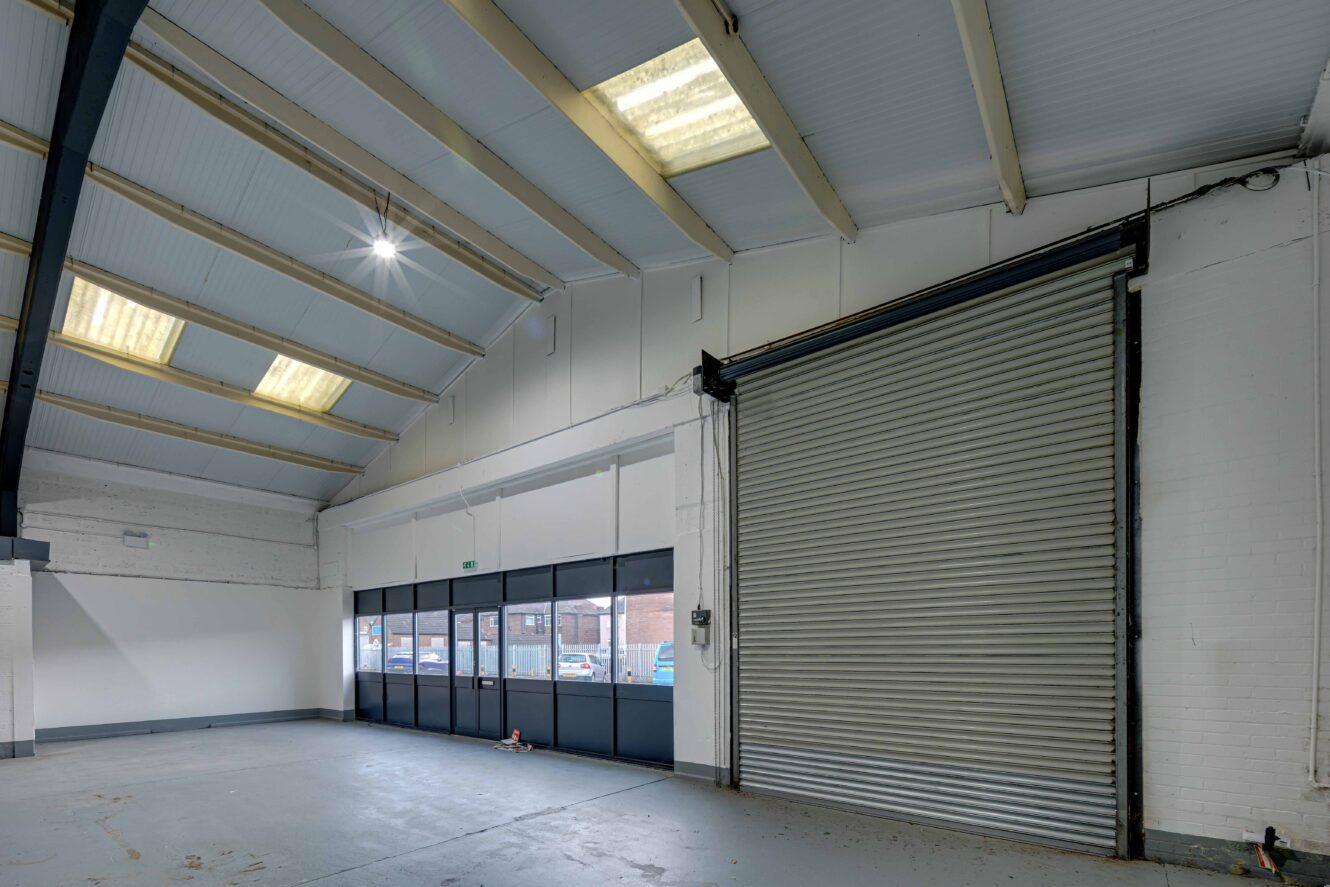 Interior of an empty industrial warehouse with a large roller shutter door, windows, and skylights letting in natural light.