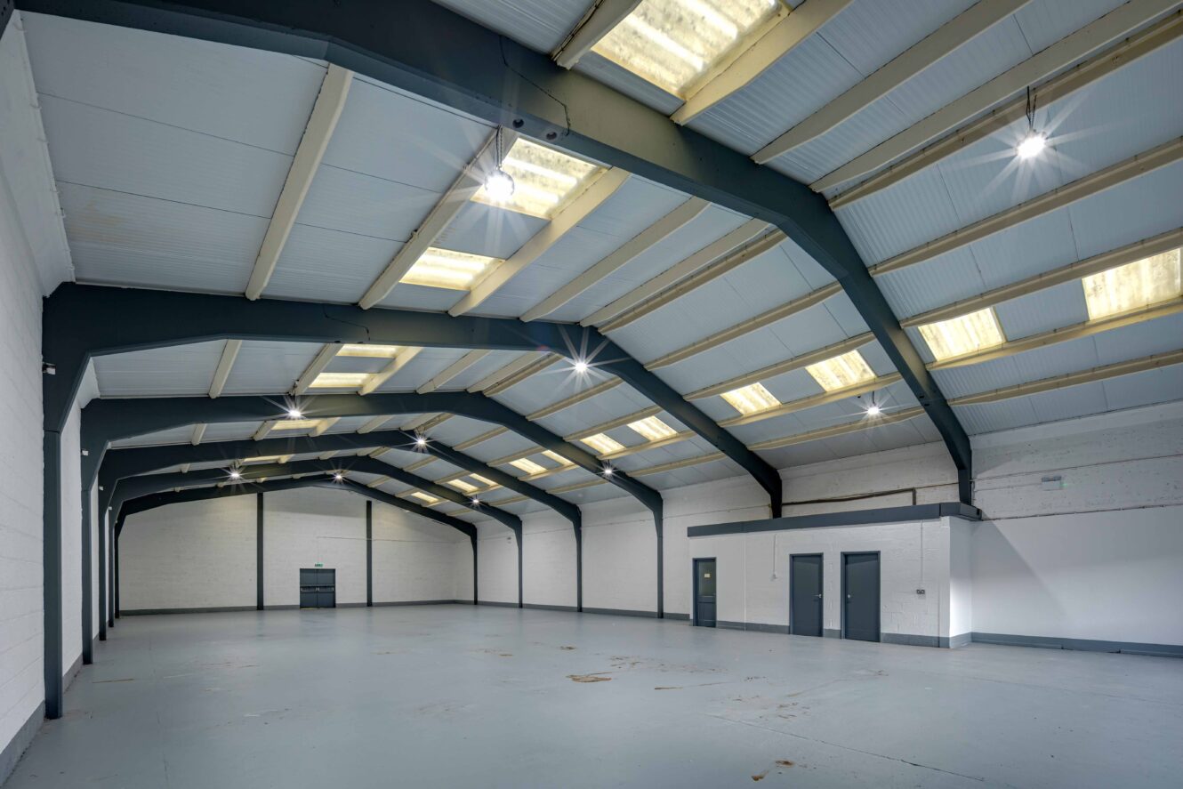 Large, empty industrial warehouse with high ceilings, skylights, gray floor, and several closed doors along the side wall.