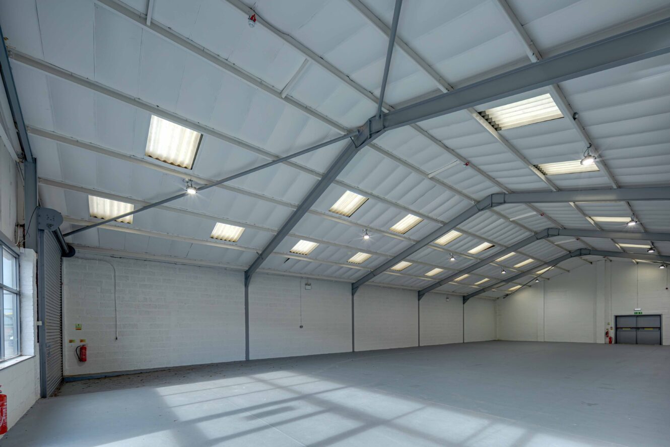 Interior of a large, empty industrial warehouse with a high ceiling, white walls, skylights, fluorescent lights, and a concrete floor.