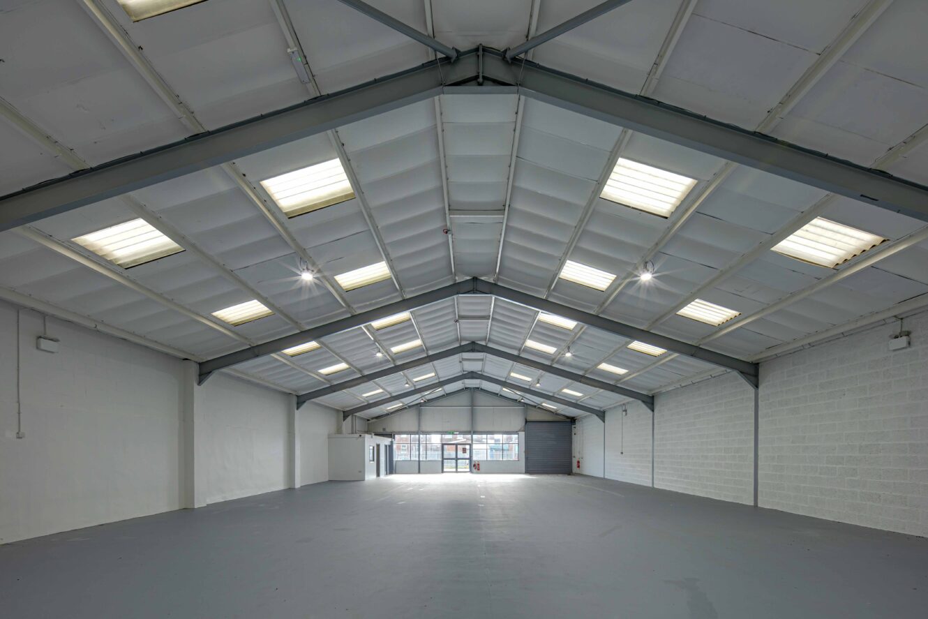 Interior of an empty industrial warehouse with a high ceiling, exposed beams, skylights, and large roller doors at the far end.