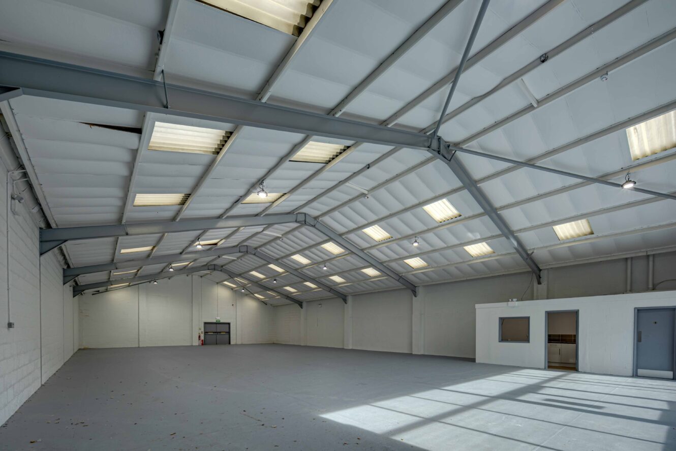 Empty industrial warehouse with high ceilings, exposed beams, large overhead lighting, white walls, and a small office area in one corner.