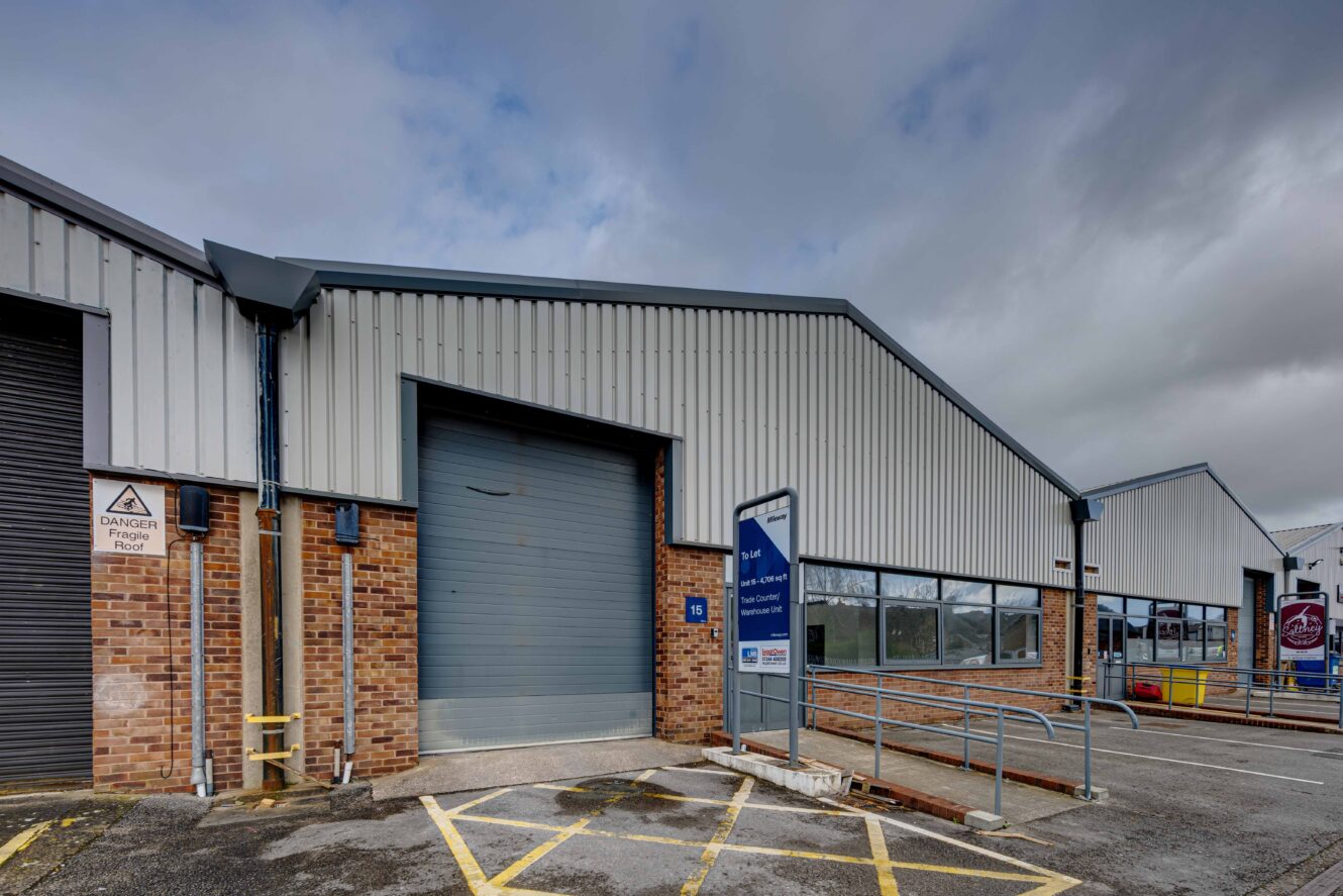 Single-story industrial building with brick and metal siding, a closed roller shutter door, parking spaces, and a sign near an accessibility ramp. Cloudy sky overhead.