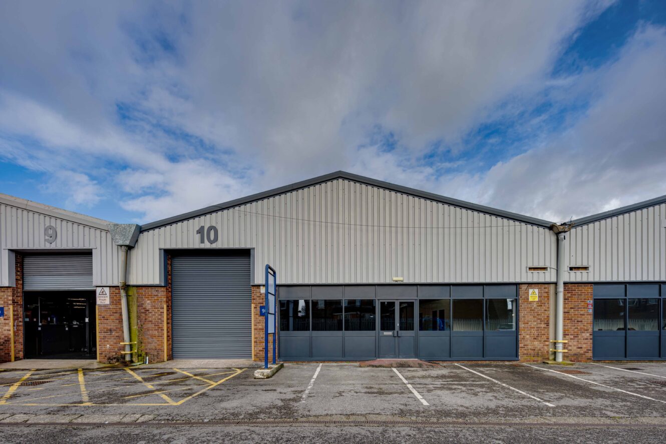 Industrial warehouse unit number 10 with a large roller shutter door, adjacent parking spaces, and a partly cloudy sky overhead.