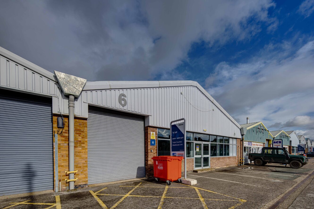 Industrial building with the number 6, metal shutters, a red bin, and a parked vehicle outside under a partly cloudy sky.