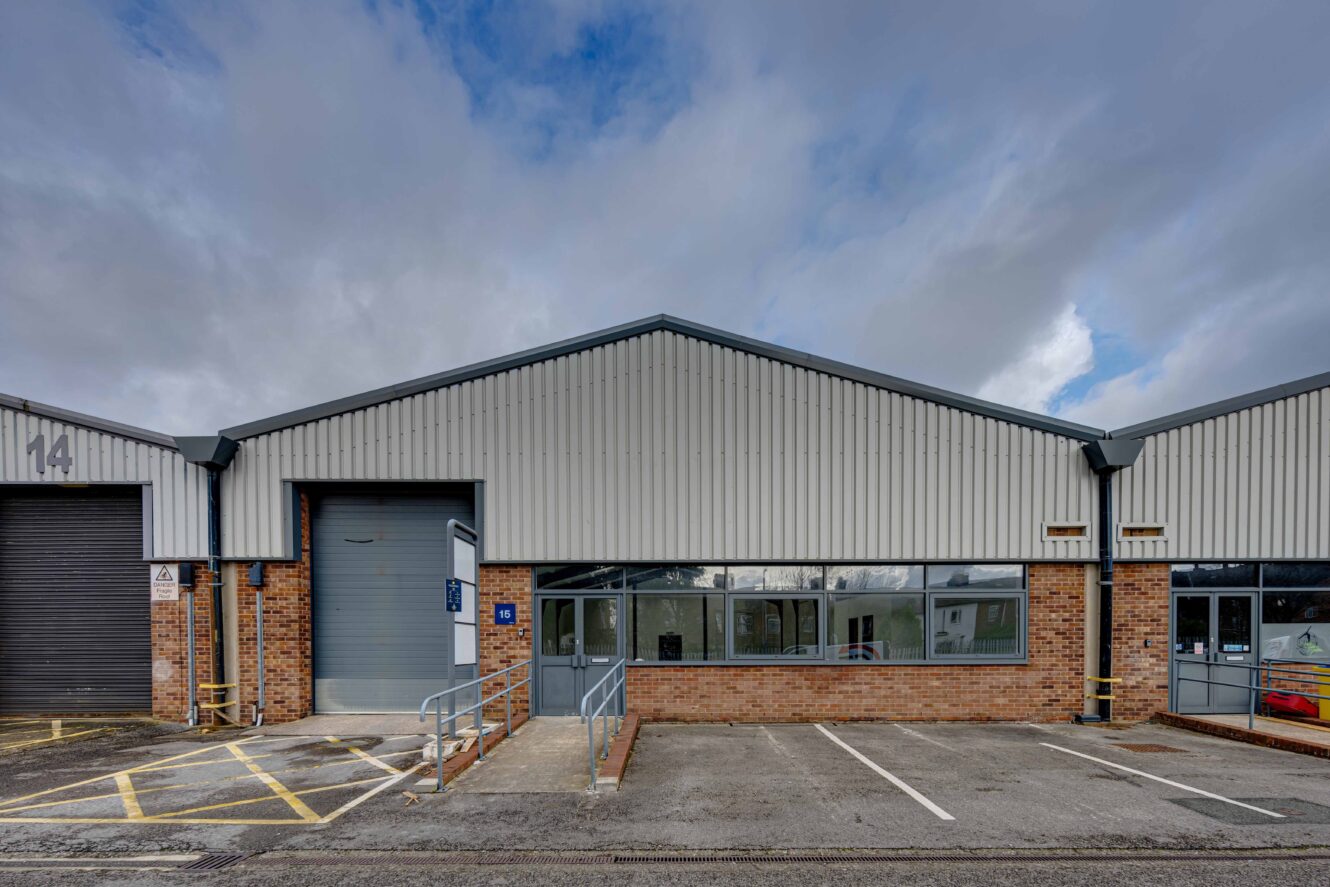 A single-story industrial warehouse with a roller shutter door, brick and metal exterior, ramp access, and adjacent parking spaces under a cloudy sky.