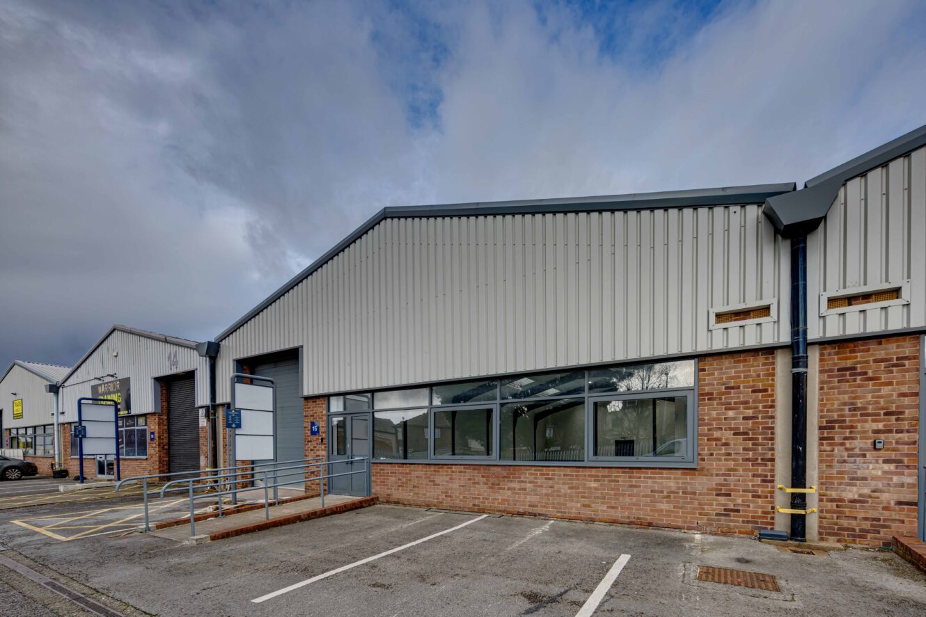 A single-story industrial building with brick and metal siding, large windows, wheelchair ramp, and empty parking spaces in front under a cloudy sky.