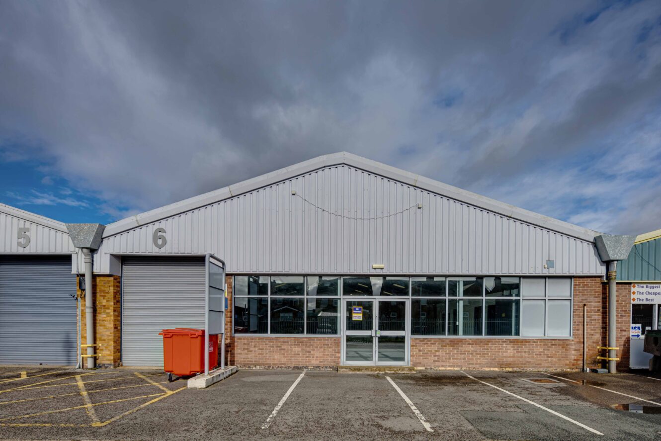A gray industrial warehouse building with glass entrance doors, large windows, and numbered units 5 and 6. There is a red bin and parking area in front under a cloudy sky.