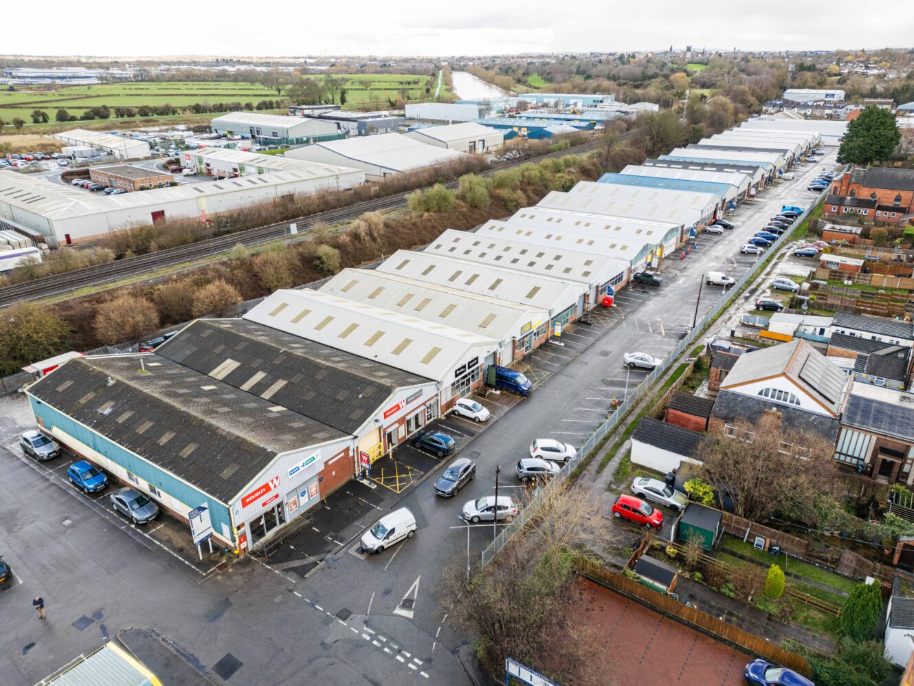 Aerial view of an industrial estate with rows of warehouses, parked cars, and surrounding fields under a cloudy sky.