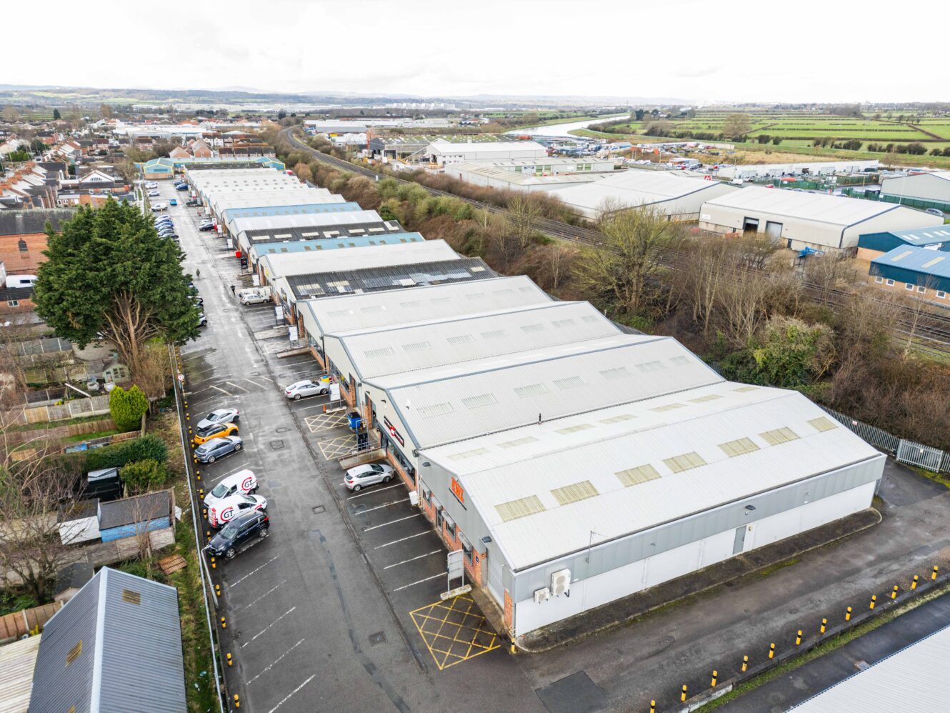 Aerial view of industrial warehouses with parked vehicles along access roads, surrounded by trees and adjacent fields under a cloudy sky.