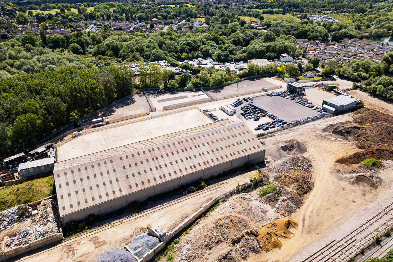 Aerial view of a large industrial warehouse with adjacent parking lot, construction materials, dirt areas, and surrounding greenery near a residential neighborhood.