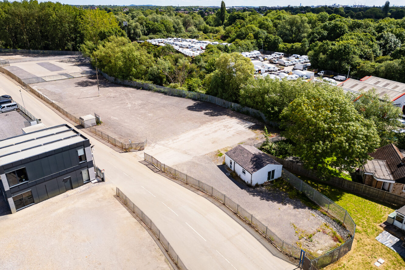 A small white building sits by a fenced, empty lot near a road, surrounded by trees and adjacent to a large area filled with parked vehicles and trailers.