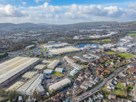Aerial view of an industrial estate with warehouses and nearby residential houses, set in a valley with hills and a town in the background under a partly cloudy sky.