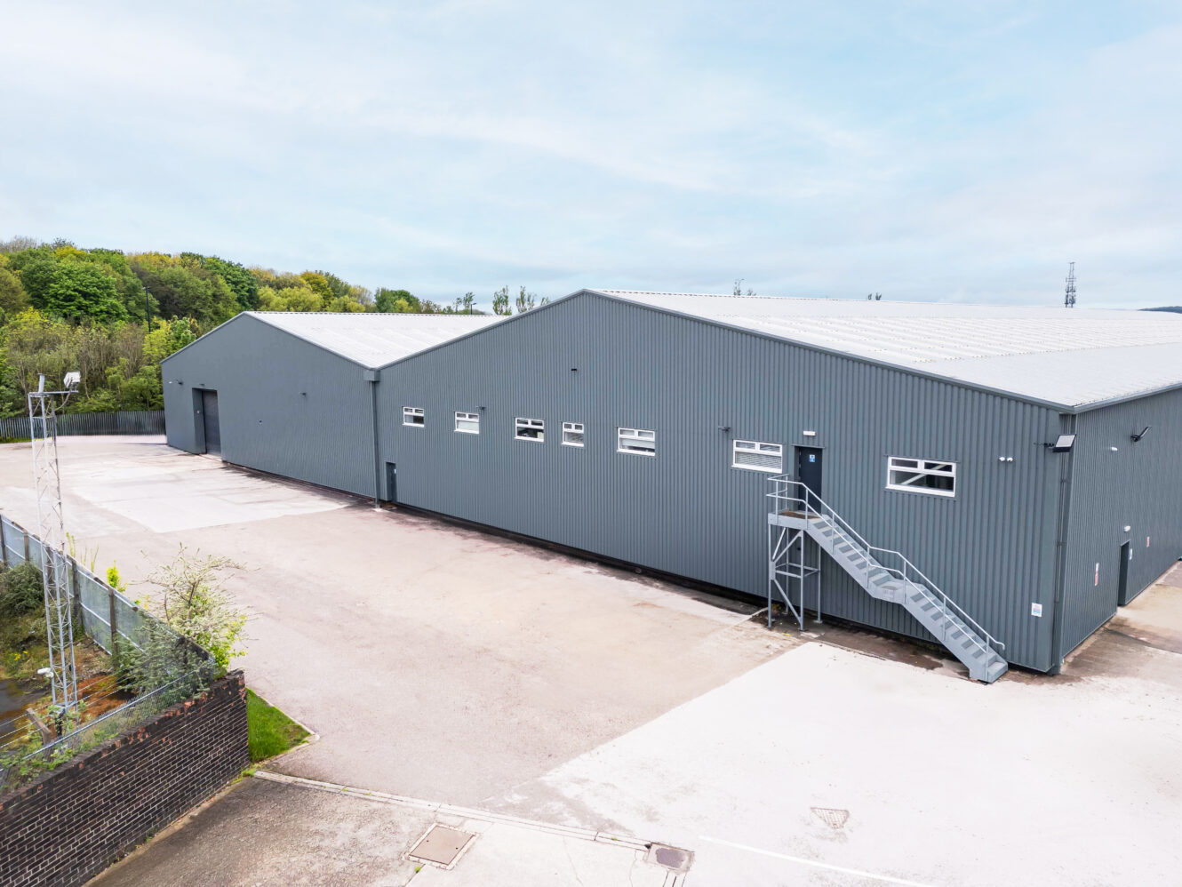 Large gray industrial warehouse with metal siding, multiple windows, and an exterior staircase, surrounded by a paved lot and some greenery in the background.