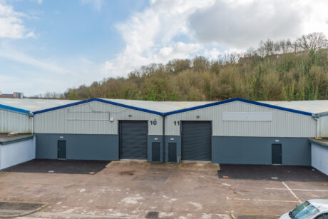 Two industrial warehouse units with large shutter doors, labeled 10 and 11, are shown with empty parking spaces in front and trees in the background.