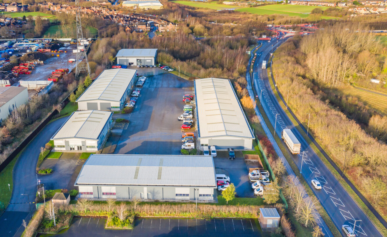 Aerial view of an industrial estate with multiple warehouses, parked vehicles, and adjacent roadways bordered by trees and grassy areas.