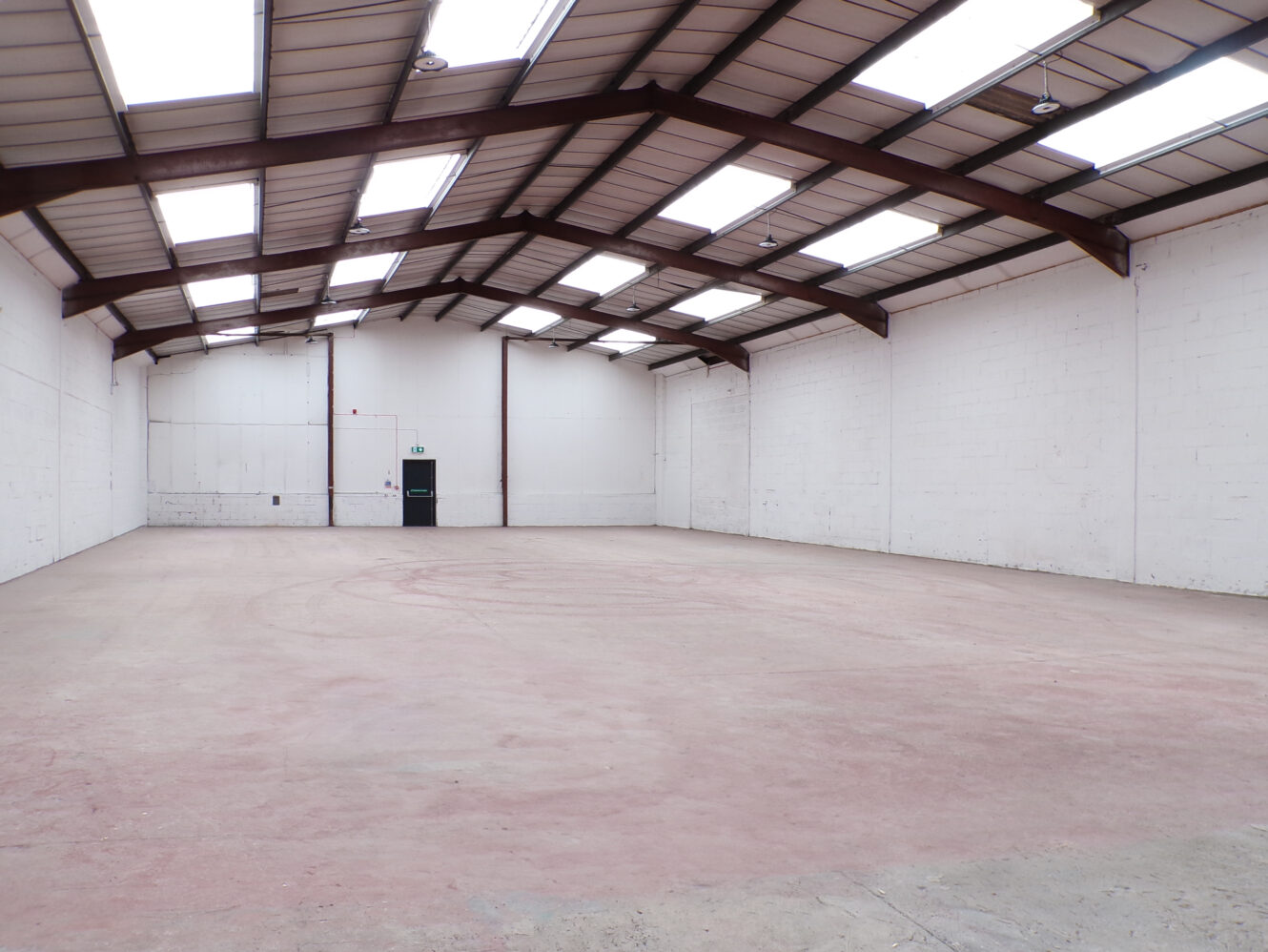 Empty warehouse interior with high ceilings, white painted brick walls, skylights, and a concrete floor; a single green door is visible at the far end.