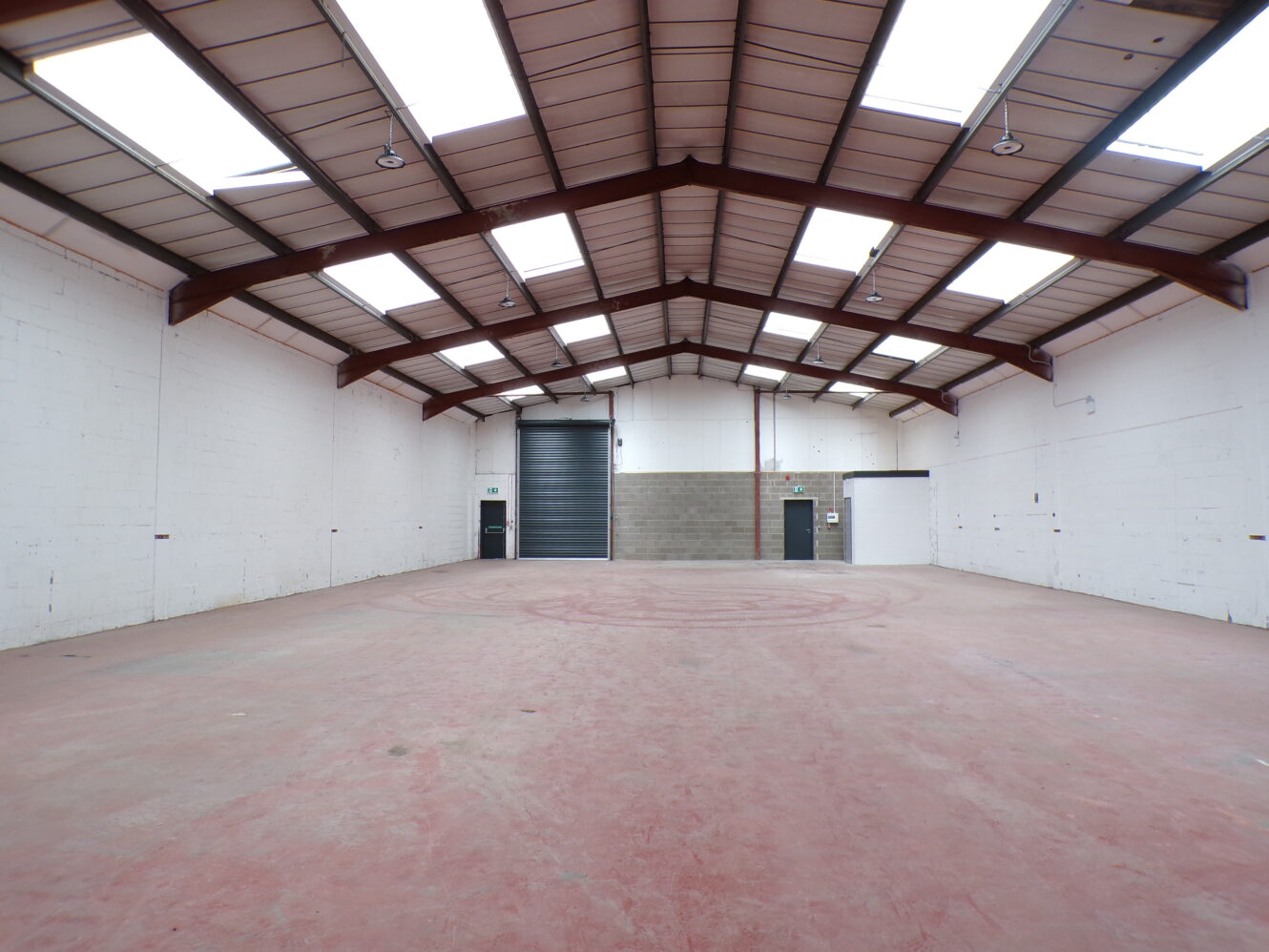 Wide-angle view of an empty industrial warehouse with red concrete floor, white walls, high ceiling, skylights, and two doors at the far end.