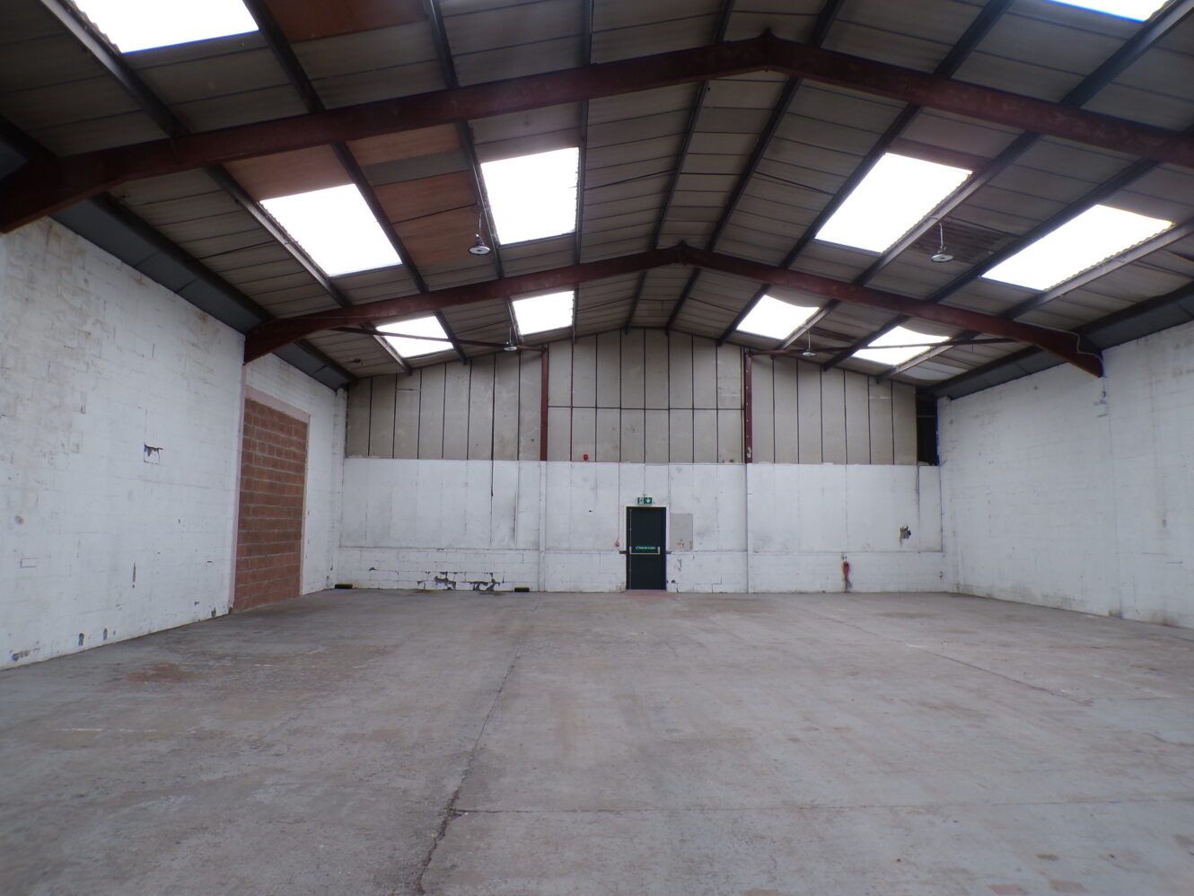 Empty warehouse interior with concrete floor, white walls, high ceiling, and skylights. A single door is visible at the far end of the space.