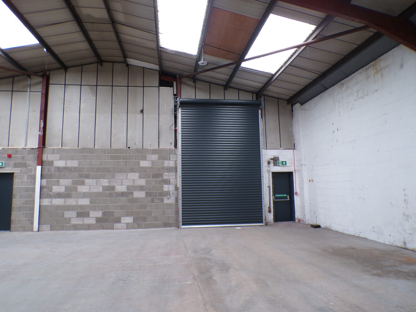 Interior of an empty warehouse with a partially open roller shutter door, concrete floor, and exposed beams on the ceiling.