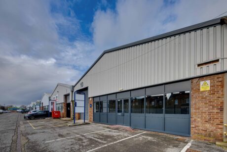 A row of industrial warehouse units with metal roofs and brick walls; one unit is labeled 10 and has large windows and roller doors, with a mostly empty parking lot in front.