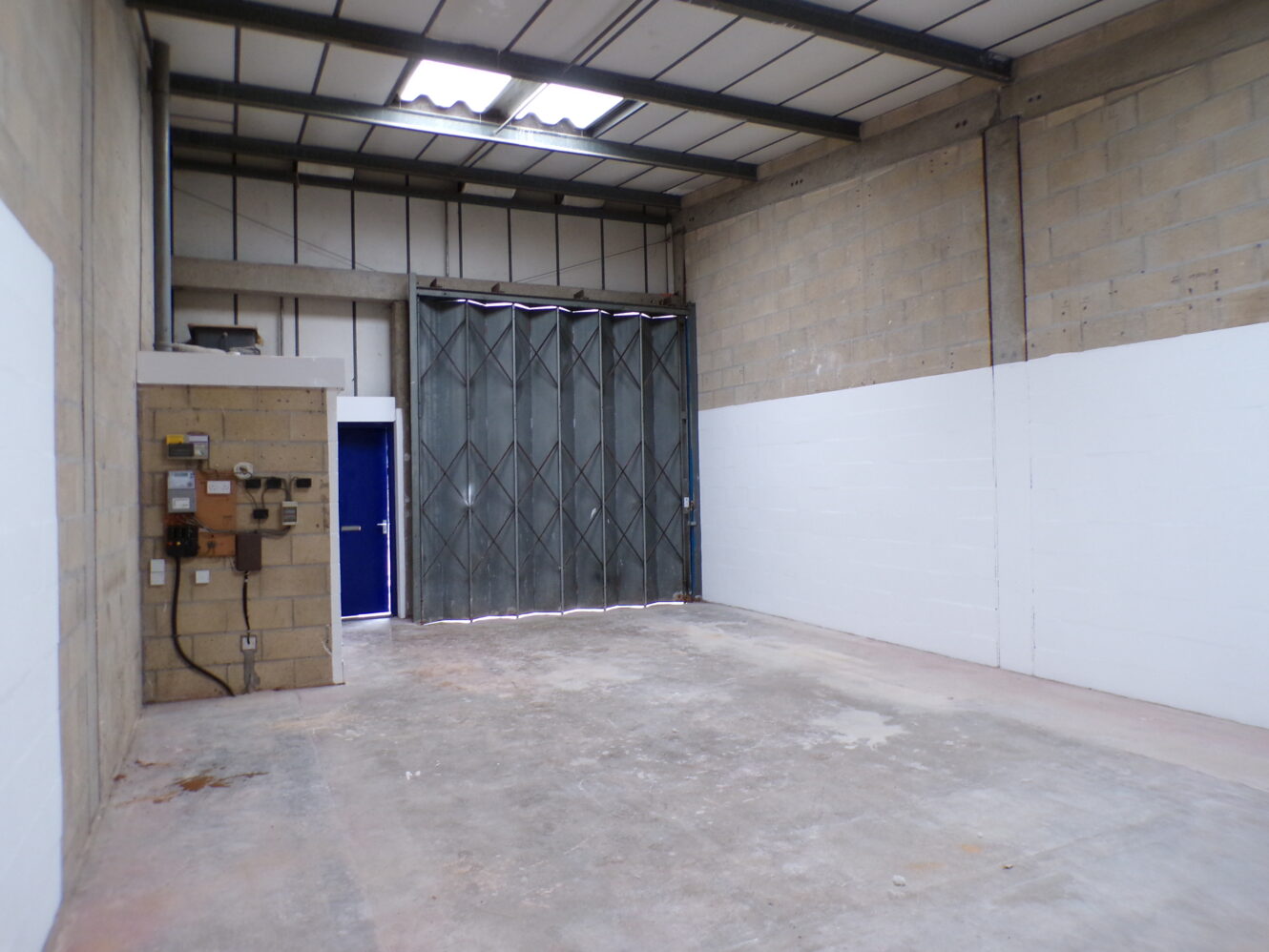 Empty industrial unit with concrete floor, high ceiling, exposed wiring, and a large metal folding door at the far end.