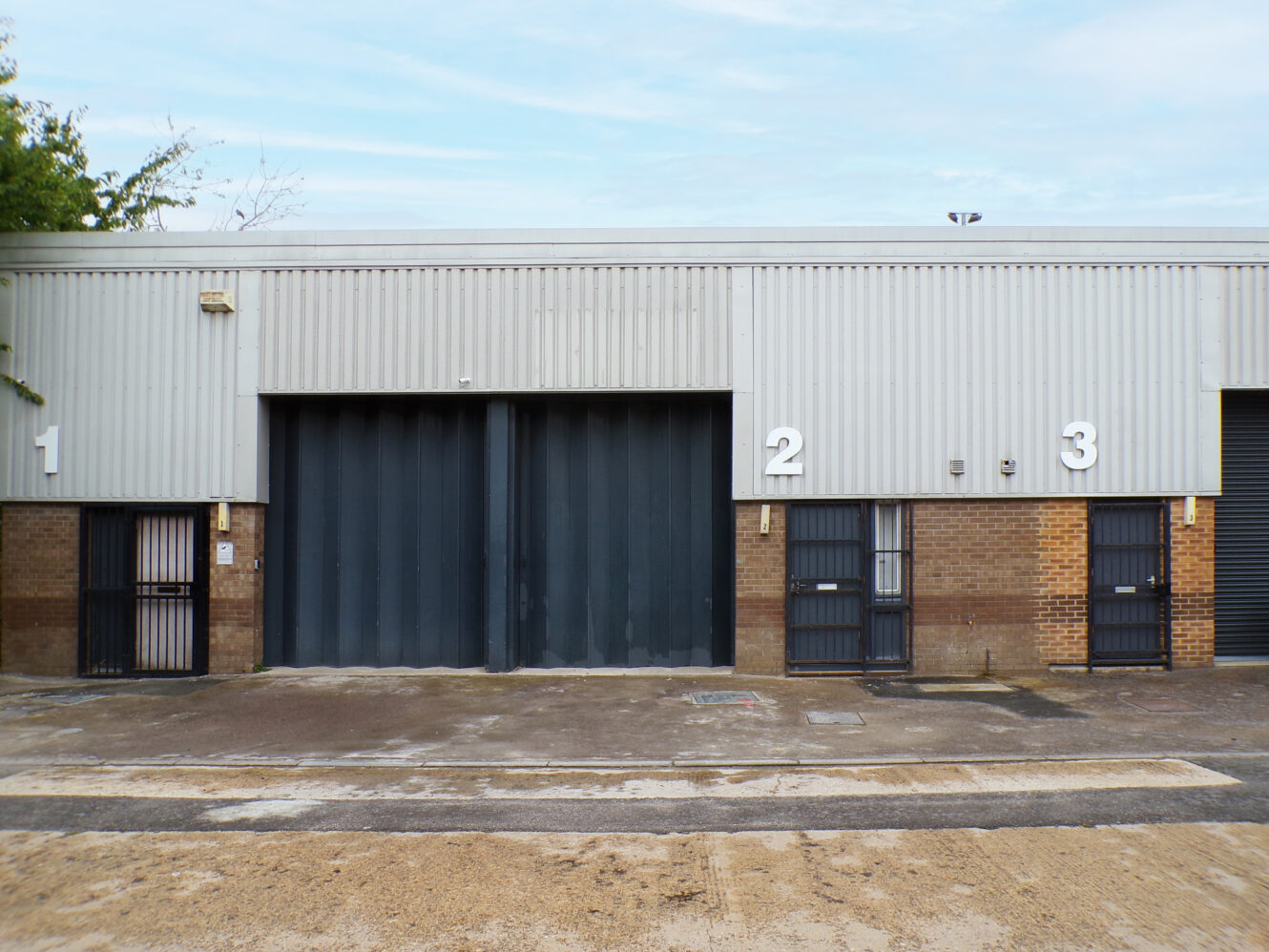 A row of three industrial warehouse units, numbered 1, 2, and 3, with metal doors and a concrete forecourt.