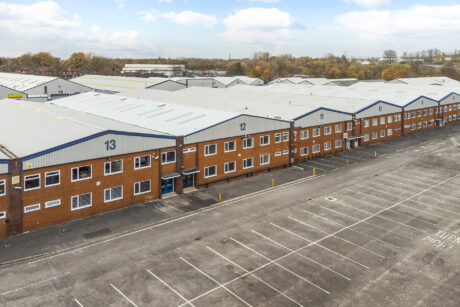 Large industrial warehouse buildings with numbered units and an adjacent empty parking lot under a partly cloudy sky.