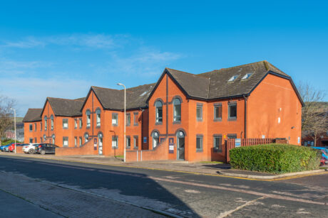 A row of modern red-brick townhouses with pitched roofs and arched windows, situated along a quiet street under a clear blue sky.