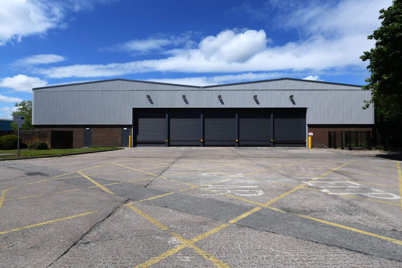 Large industrial warehouse with five closed roller doors, bordered by yellow lines in the parking area, under a blue sky with scattered clouds.