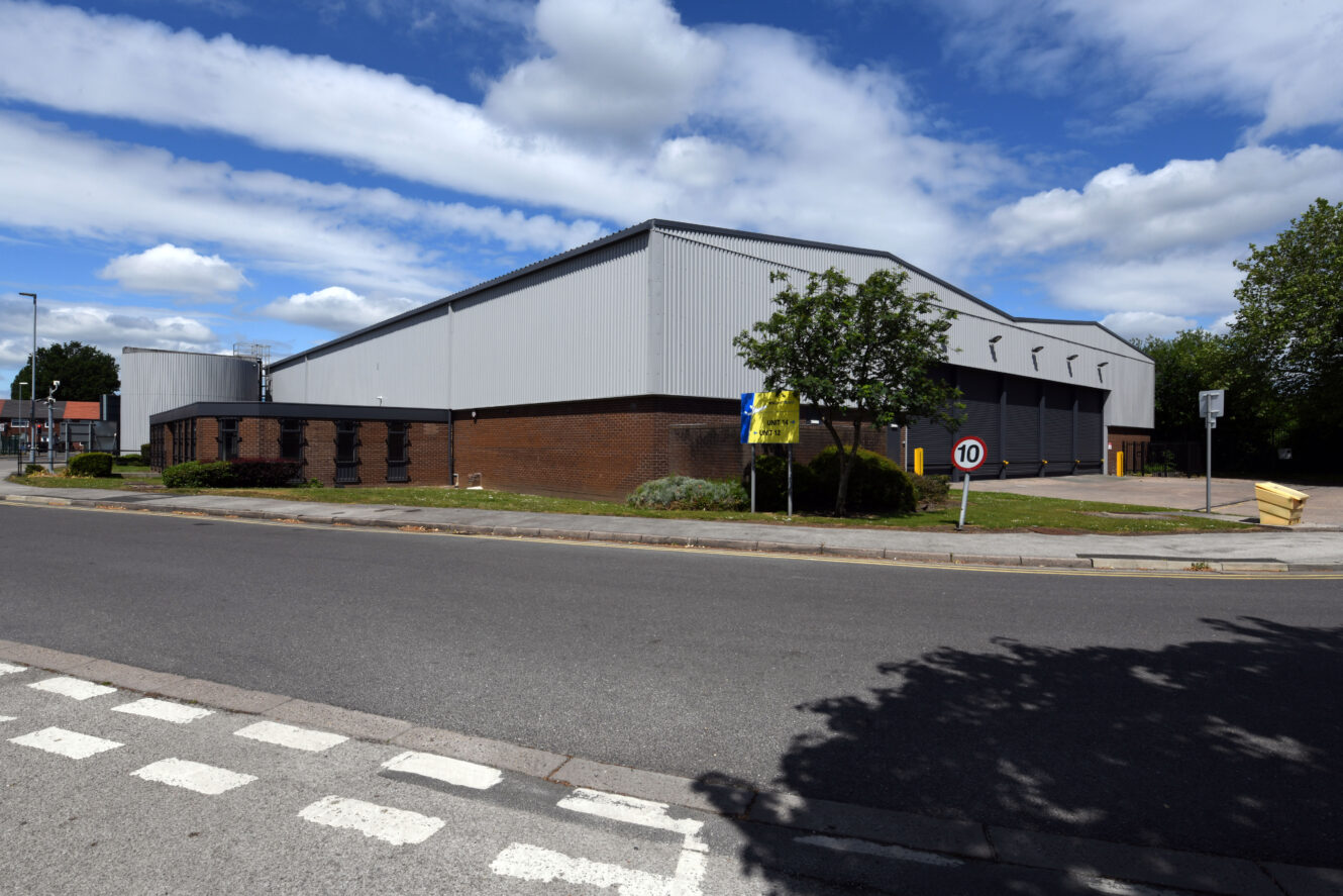 Large industrial warehouse with grey metal siding and brick base, set beside a road with a 10 mph speed limit sign and some trees under a partly cloudy sky.
