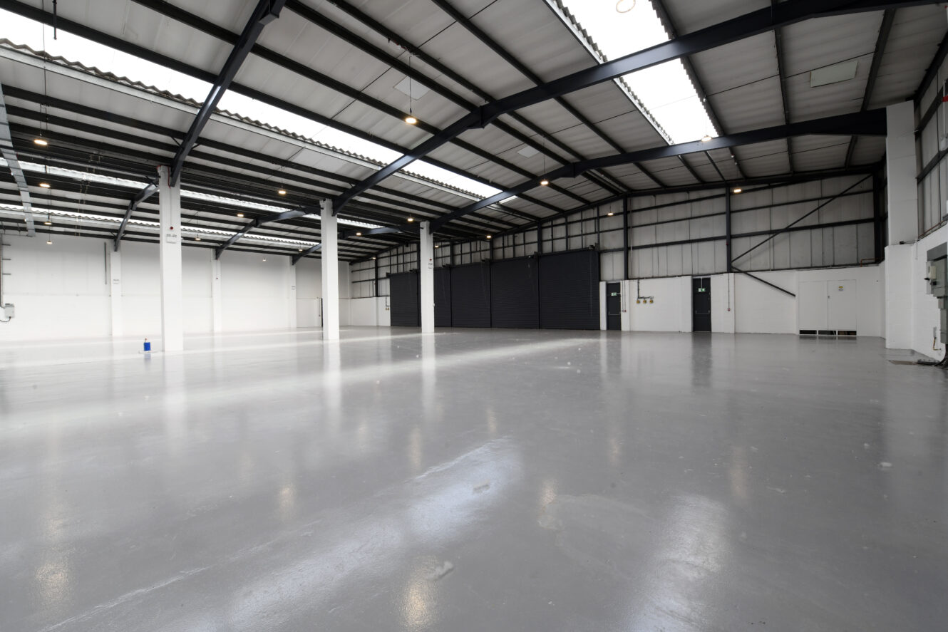 Large empty industrial warehouse with polished concrete floors, high ceilings, exposed beams, skylights, and white walls.