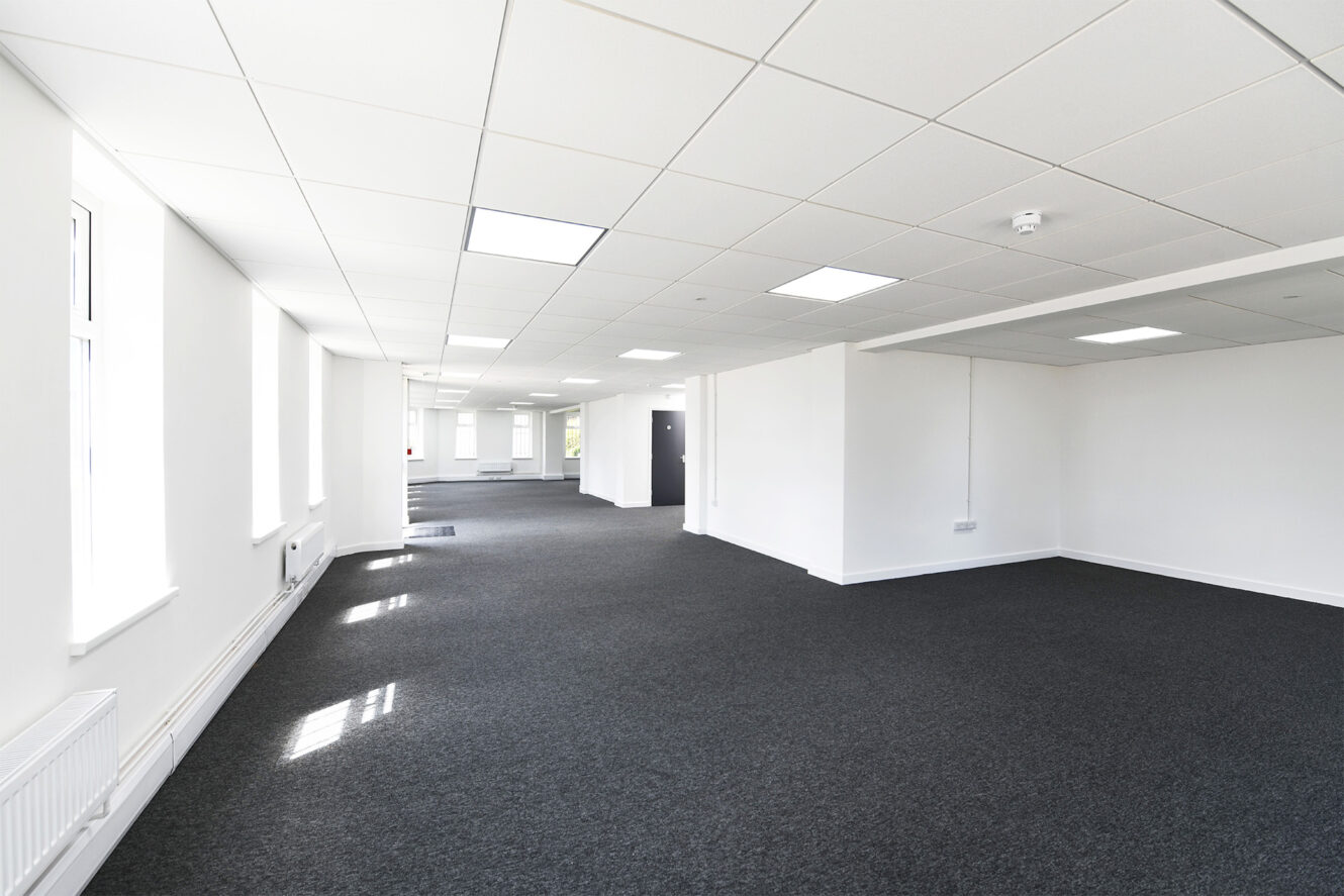 Spacious, empty office with white walls, large windows, and dark carpeted floor under a grid of ceiling lights.